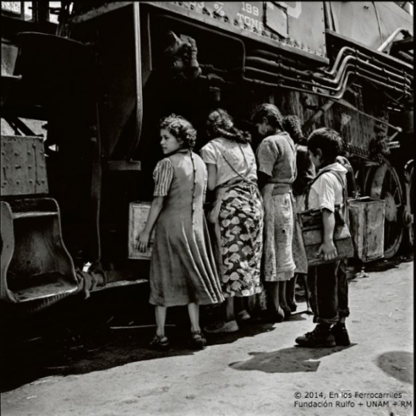 Cuatro mujeres cargan sus recipientes con agua del depósito de una locomotora de vapor, mientras un niño con su cajón de lustrabotas mira la escena en la estación Tacuba. 